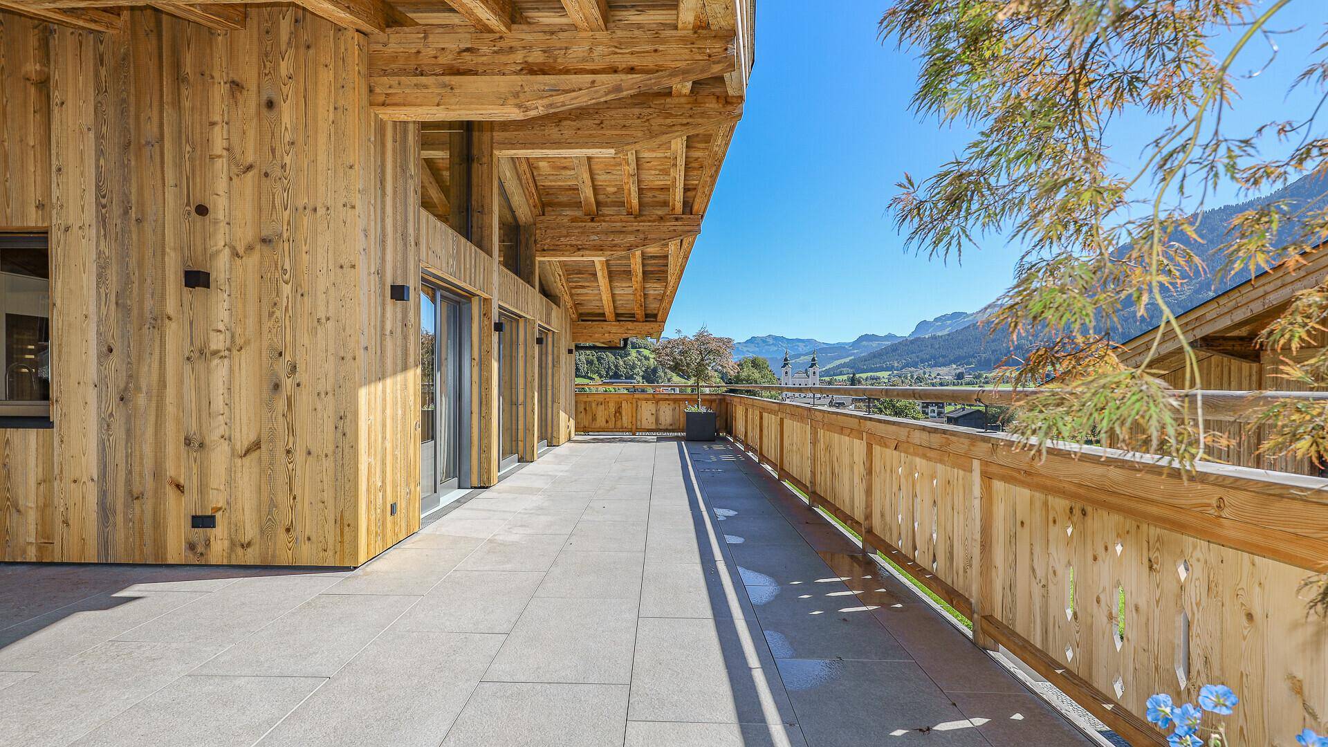 Lange Terrasse mit Holzbalkon und Steinplattenboden, bietet weite Ausblicke auf die Berglandschaft.