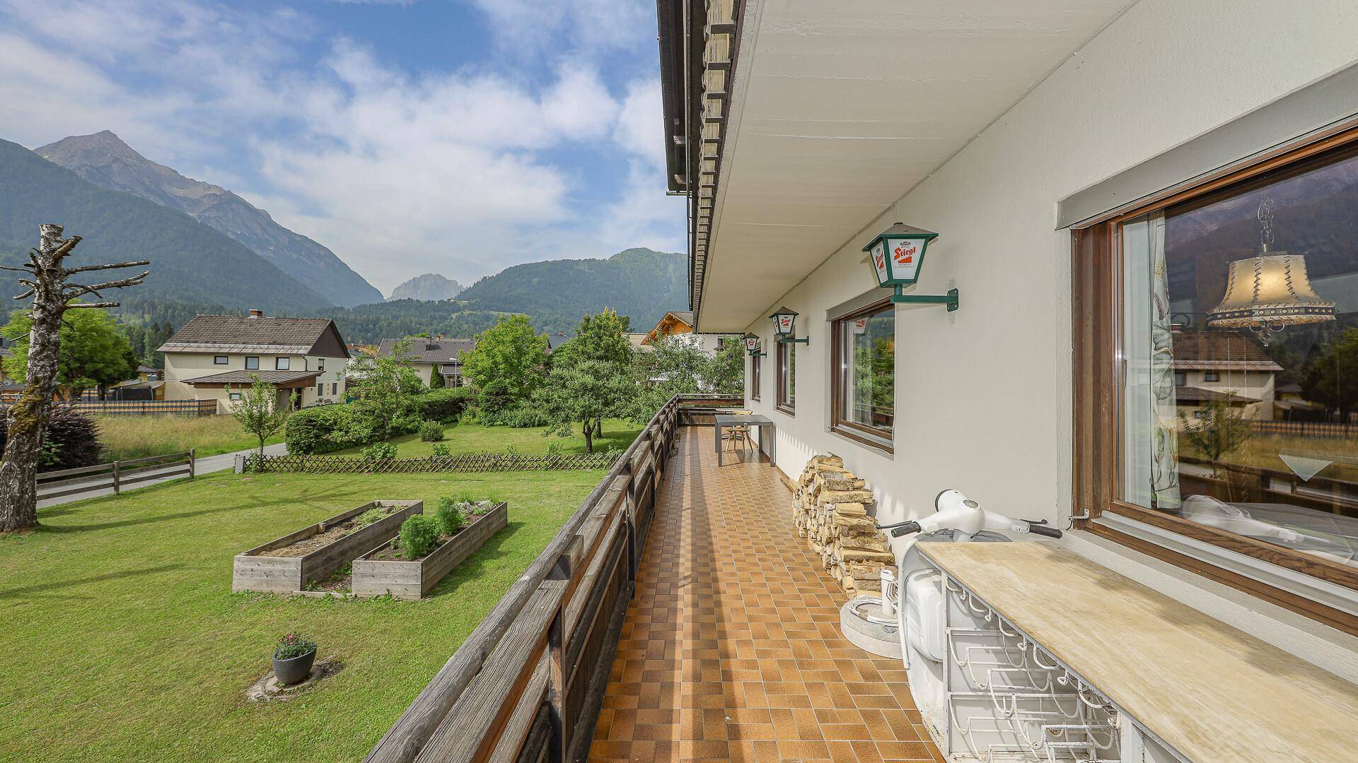 Langer Balkon mit Fliesenboden und Holzgeländer, bietet einen weiten Blick auf grüne Wiesen und Berge.