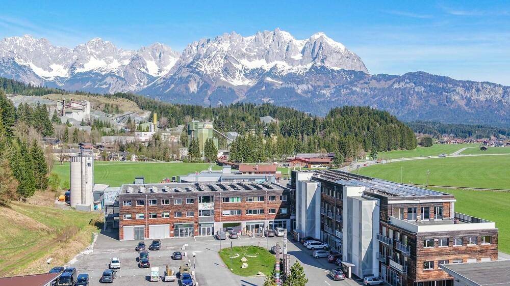 Panoramablick auf einen Gebäudekomplex mit Parkplätzen vor einer beeindruckenden Bergkulisse.