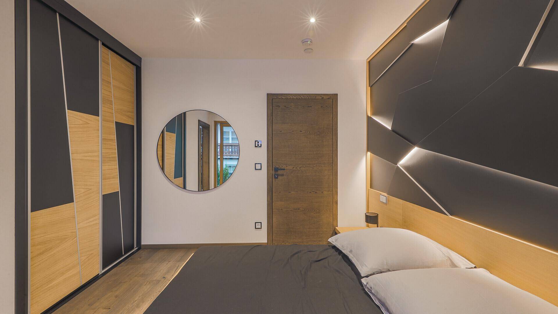 Schlafzimmer mit modernem Kopfteil, Holzboden und einem runden Spiegel an der Wand.