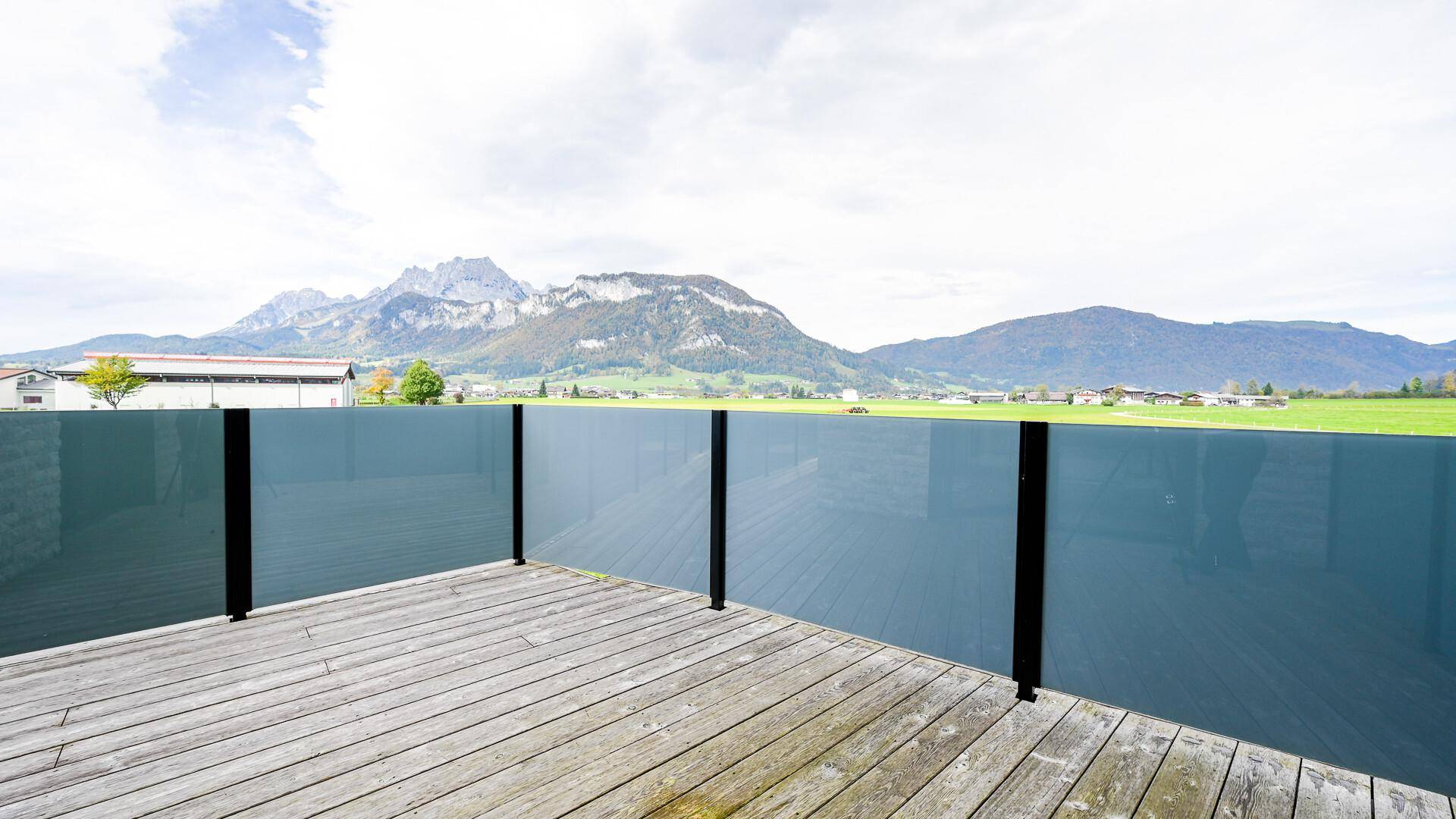 Weitläufige Holzterrasse mit Glasgeländer und beeindruckendem Blick auf die Berge und Felder.