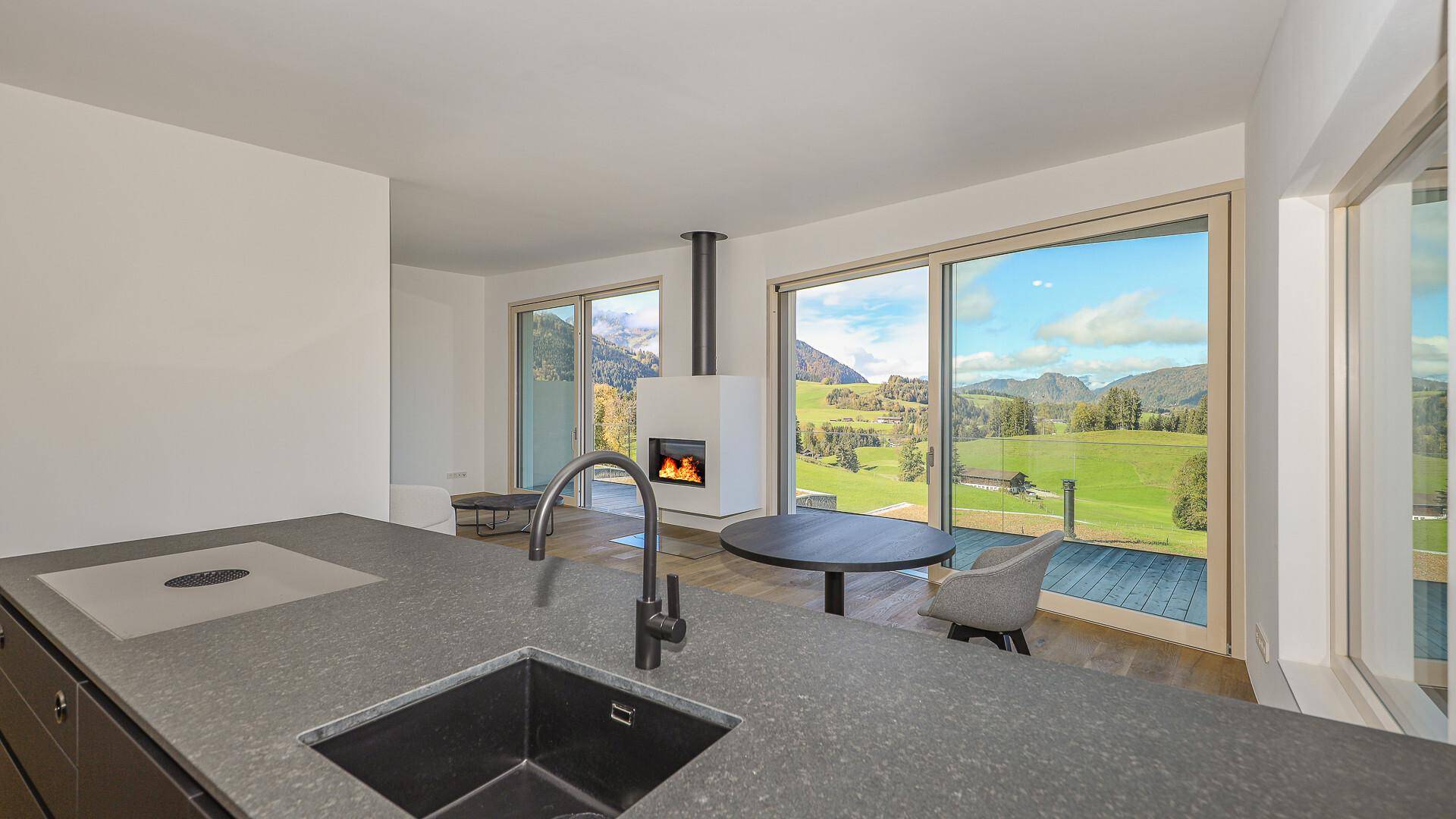 Küchenzeile mit dunkler Arbeitsplatte und Spüle, mit Blick auf den Wohnbereich und die Berge.