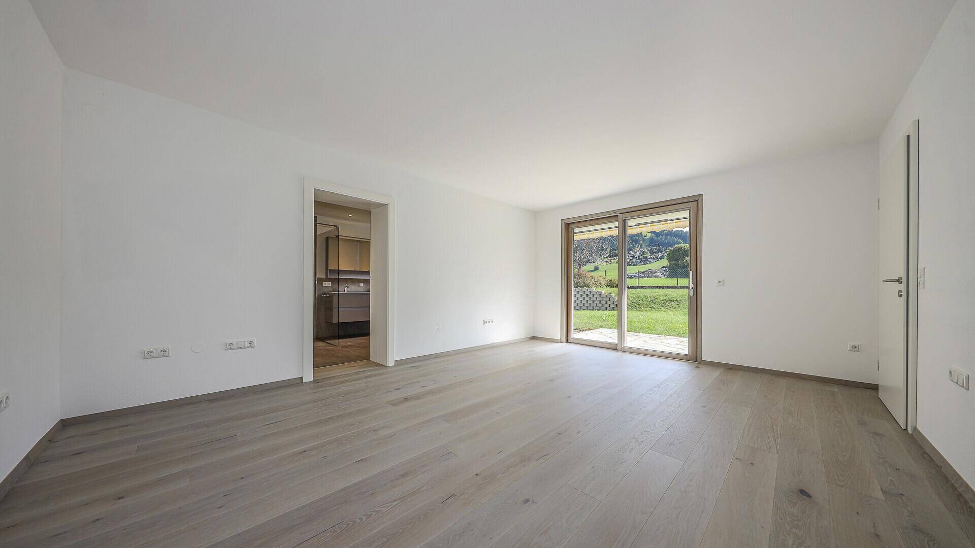 Helles, leeres Schlafzimmer mit Holzboden und großer Schiebetür zum Gartenbereich.