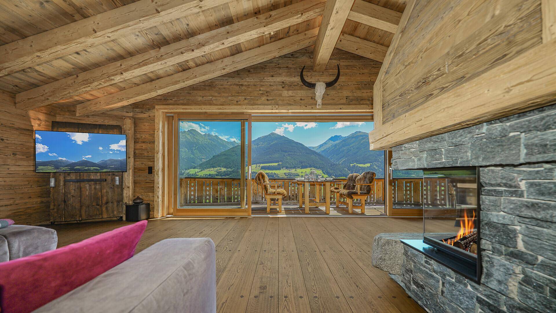 Helles Wohnzimmer mit Steinkamin, Holzbalken und raumhohen Fenstern mit Bergblick.