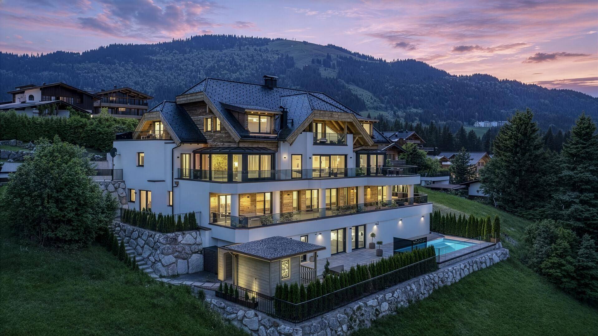 Luxuriöses Anwesen in den Bergen bei Sonnenuntergang, mit beleuchteten Balkonen und Pool.