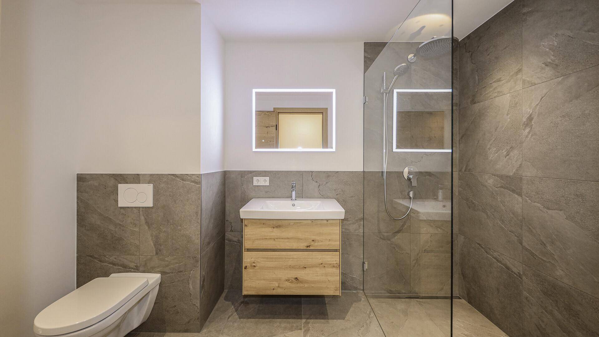 Modernes Badezimmer mit grauen Fliesen, Holzwaschtisch, wandhängendem WC und begehbarer Dusche.