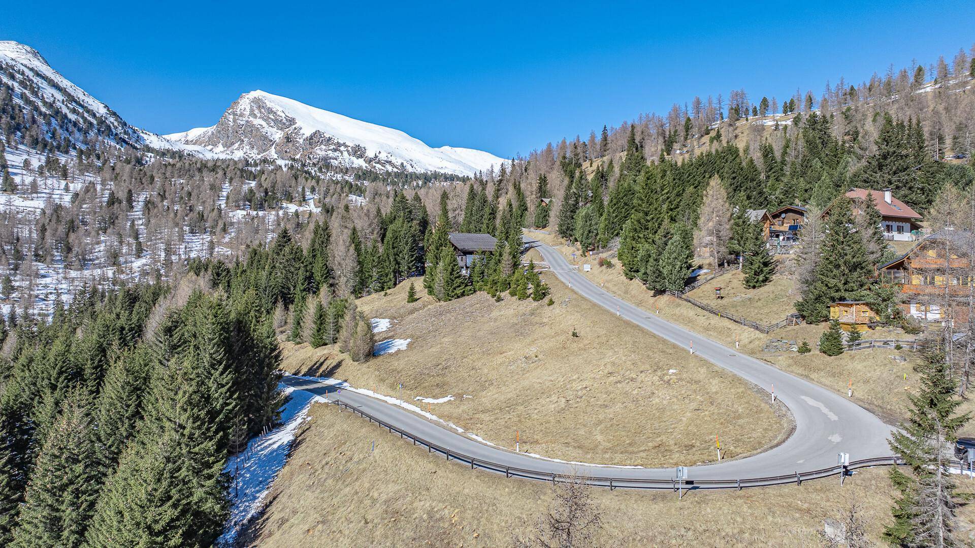 Kurvige Bergstraße schlängelt sich durch eine bewaldete Landschaft mit vereinzelten Häusern und schneebedeckten Gipfeln im Hintergrund.