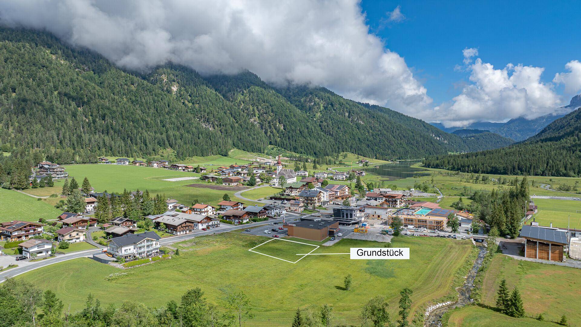 Luftaufnahme eines Dorfes in einem Tal mit Bergen, See und markiertem Grundstück für Neubau.