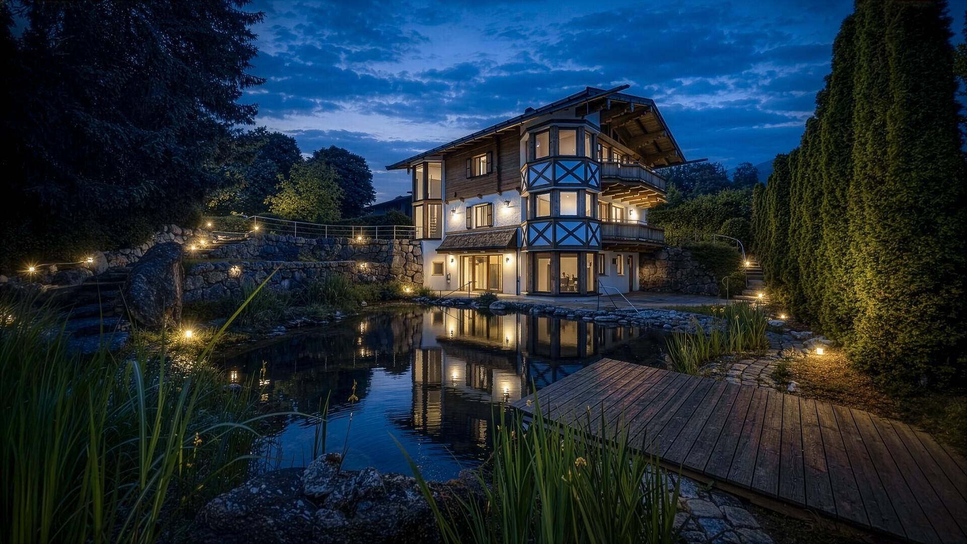 Beleuchtetes Chalet bei Nacht mit idyllischem Naturteich und Gartenbeleuchtung.