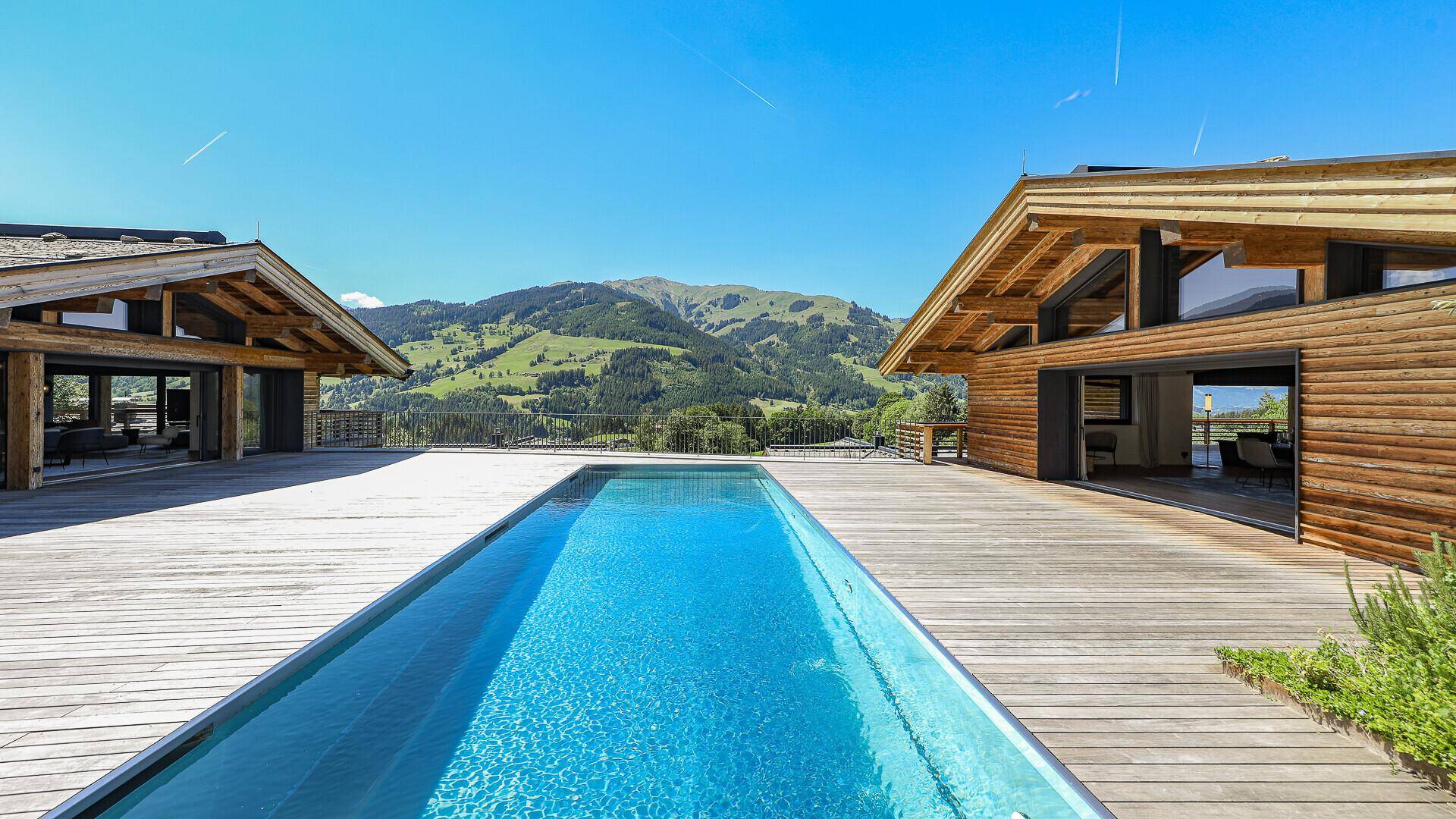 Langer Außenpool auf einer Holzterrasse zwischen zwei modernen Chalet-Gebäuden mit Bergblick.