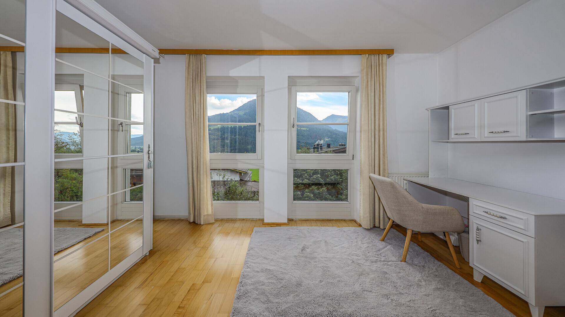 Helles Schlafzimmer mit Schreibtisch, Spiegelschrank und großen Fenstern mit Bergblick.