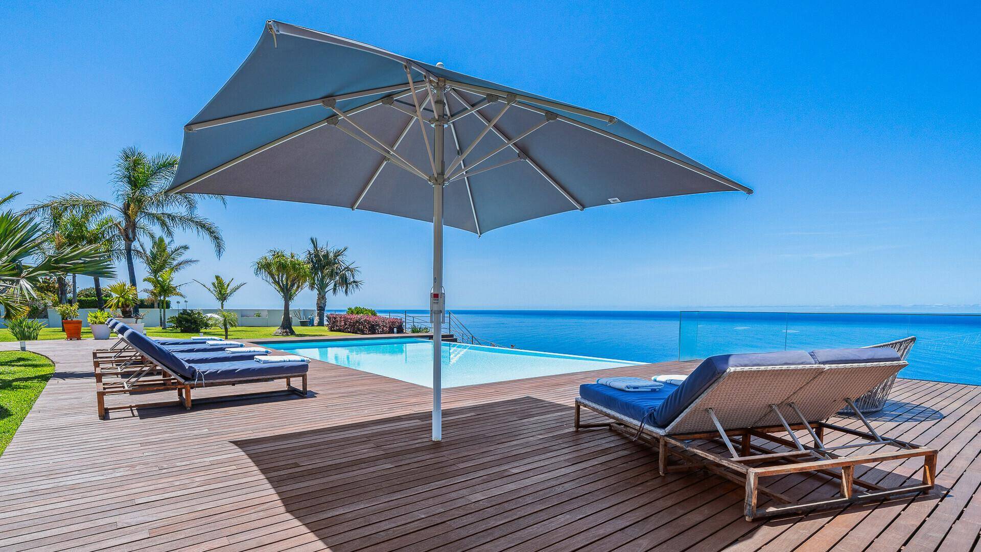 Entspannender Poolbereich mit Holzdeck, Sonnenliegen und großem Sonnenschirm, bietet einen weiten Meerblick.
