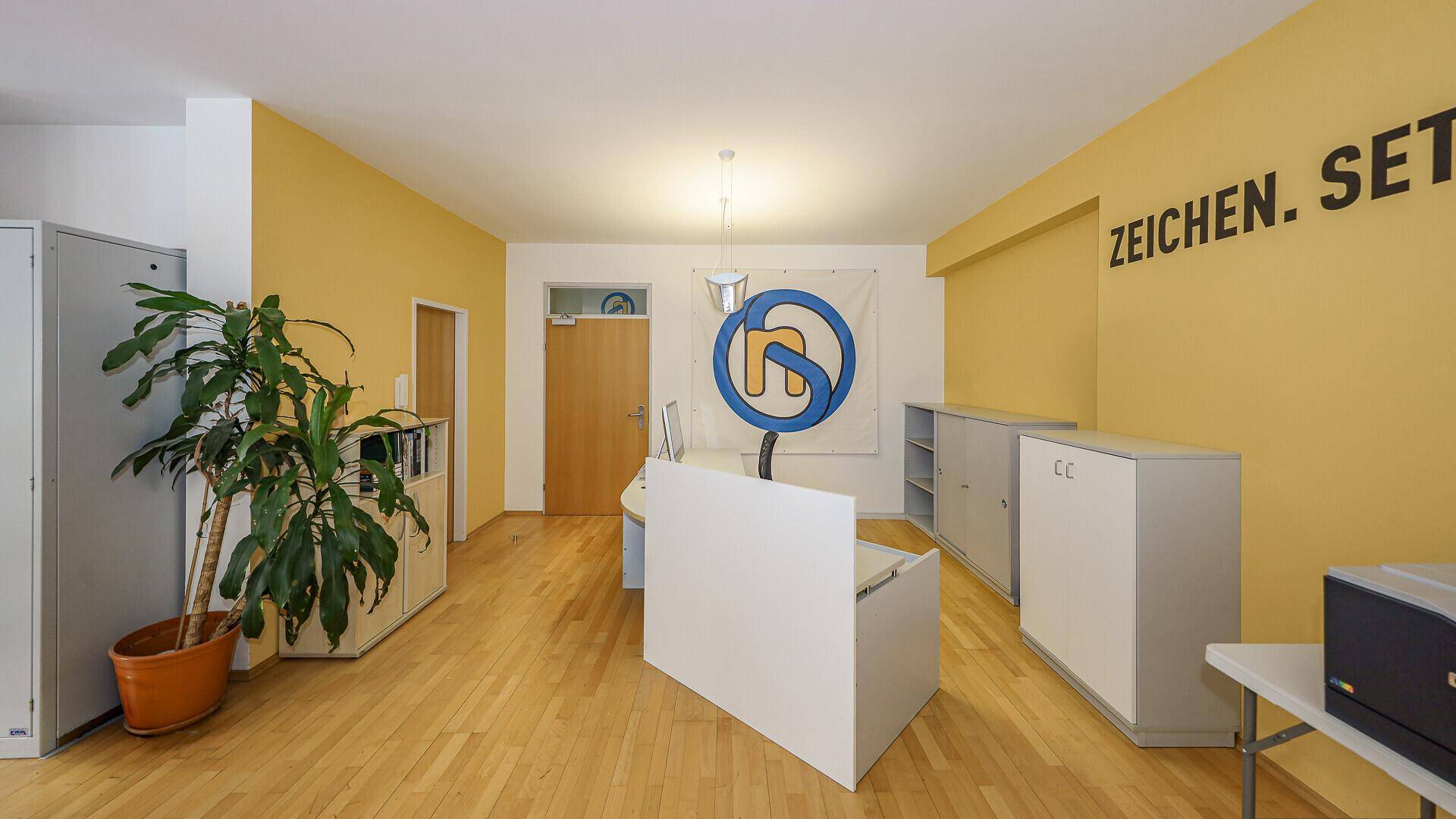 Empfangsbereich oder Büroflur mit gelber Wand, Holzboden und einer großen Pflanze, funktional eingerichtet.