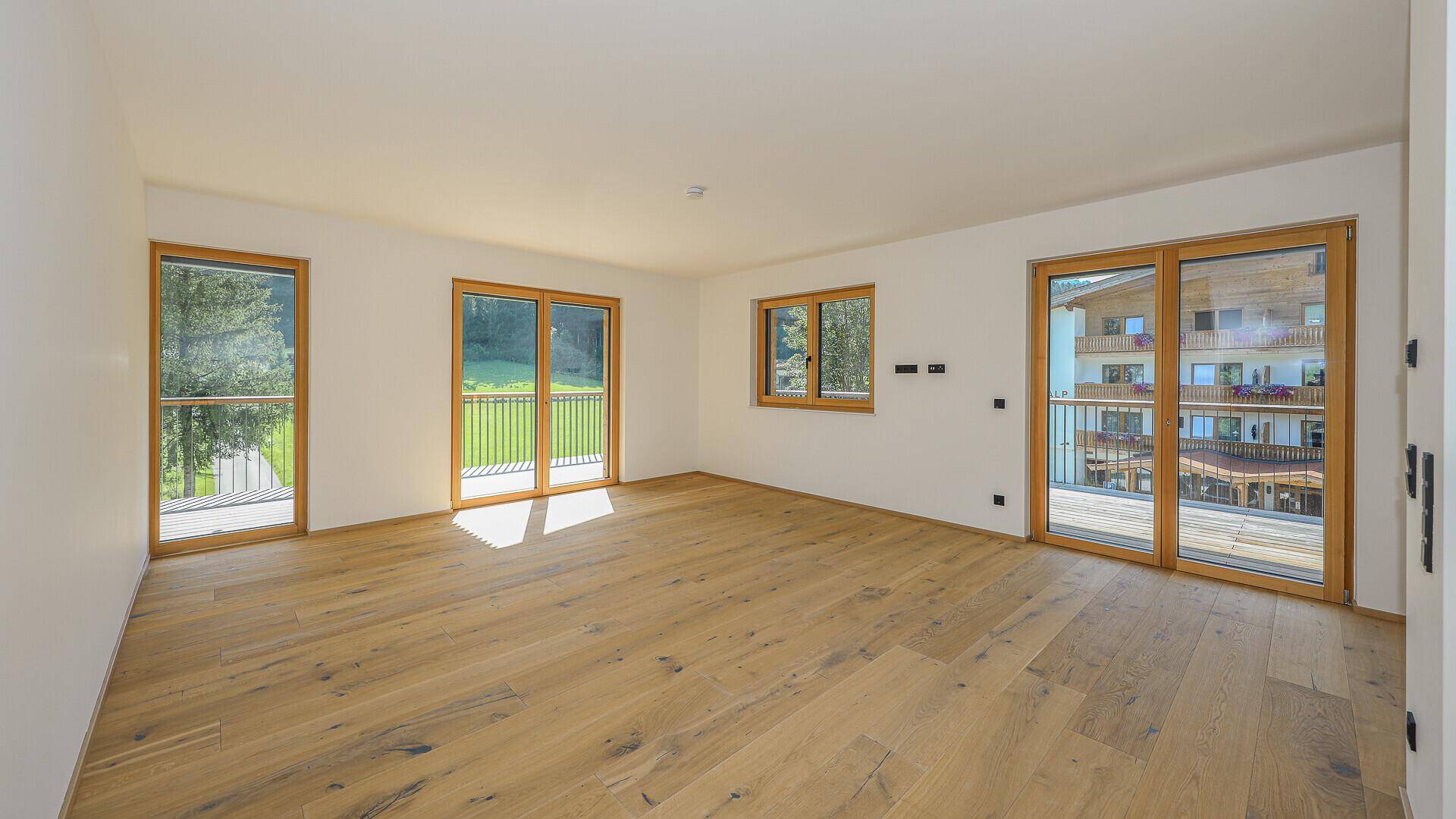 Geräumiger, heller Wohnbereich mit Holzboden und mehreren Fenstern mit Blick ins Grüne.
