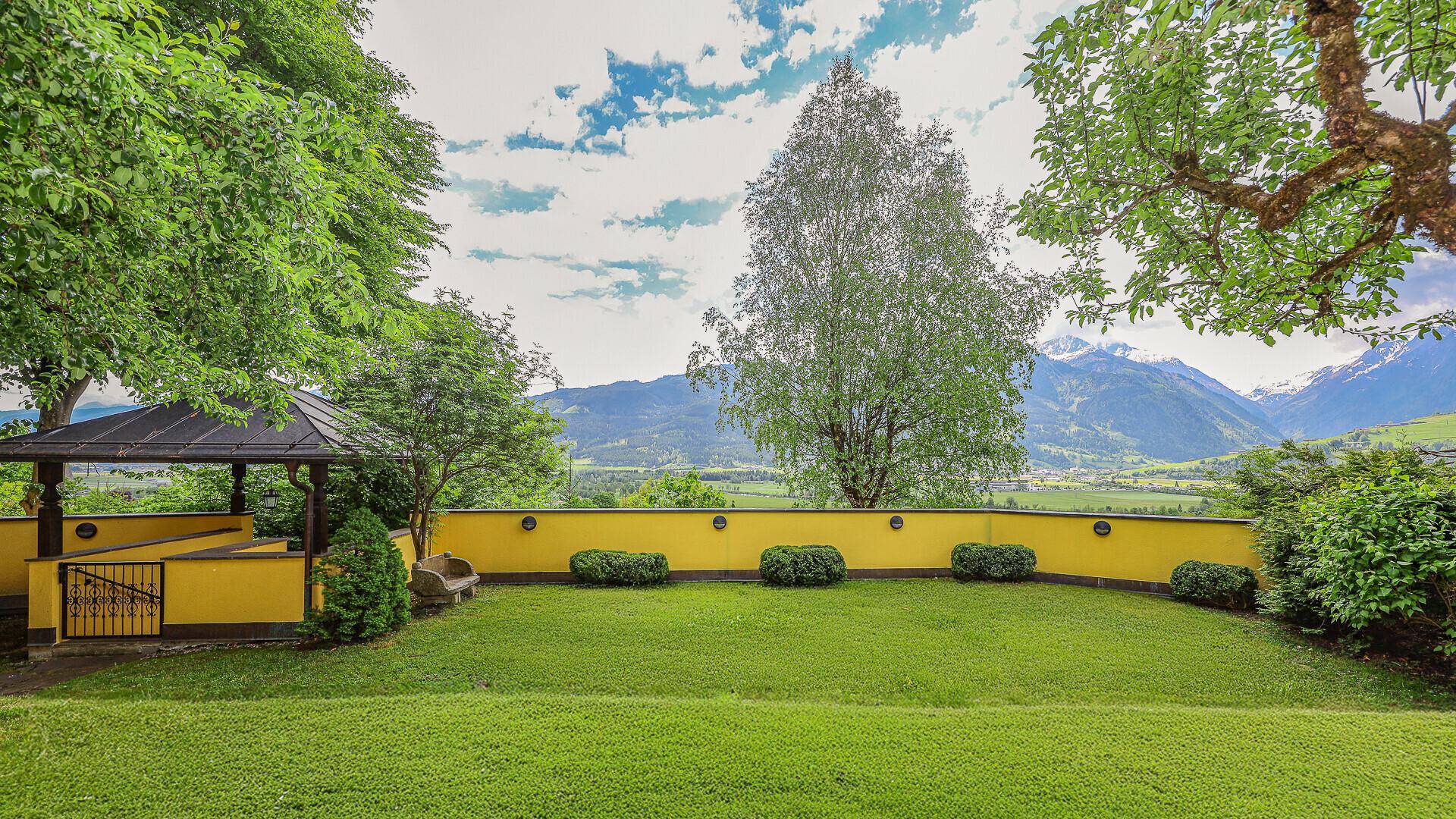 Gepflegter Gartenbereich mit Rasenfläche, Gartenlaube und malerischem Bergblick unter blauem Himmel.
