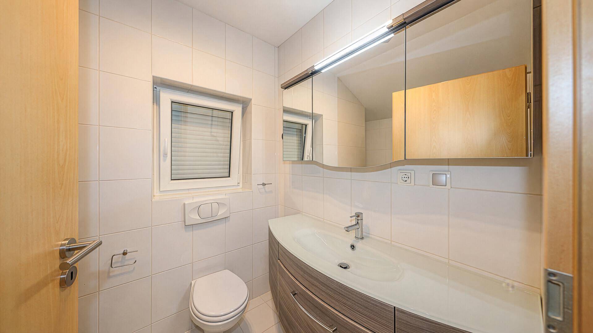 Helles Badezimmer mit WC, Waschbecken, Spiegelschrank und Fenster für Tageslicht.
