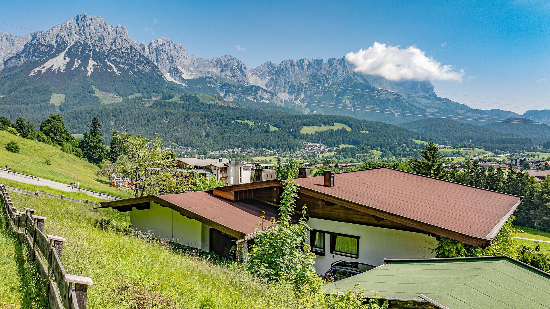 Traditionelles Haus mit braunem Dach und grüner Umgebung vor majestätischem Bergpanorama.