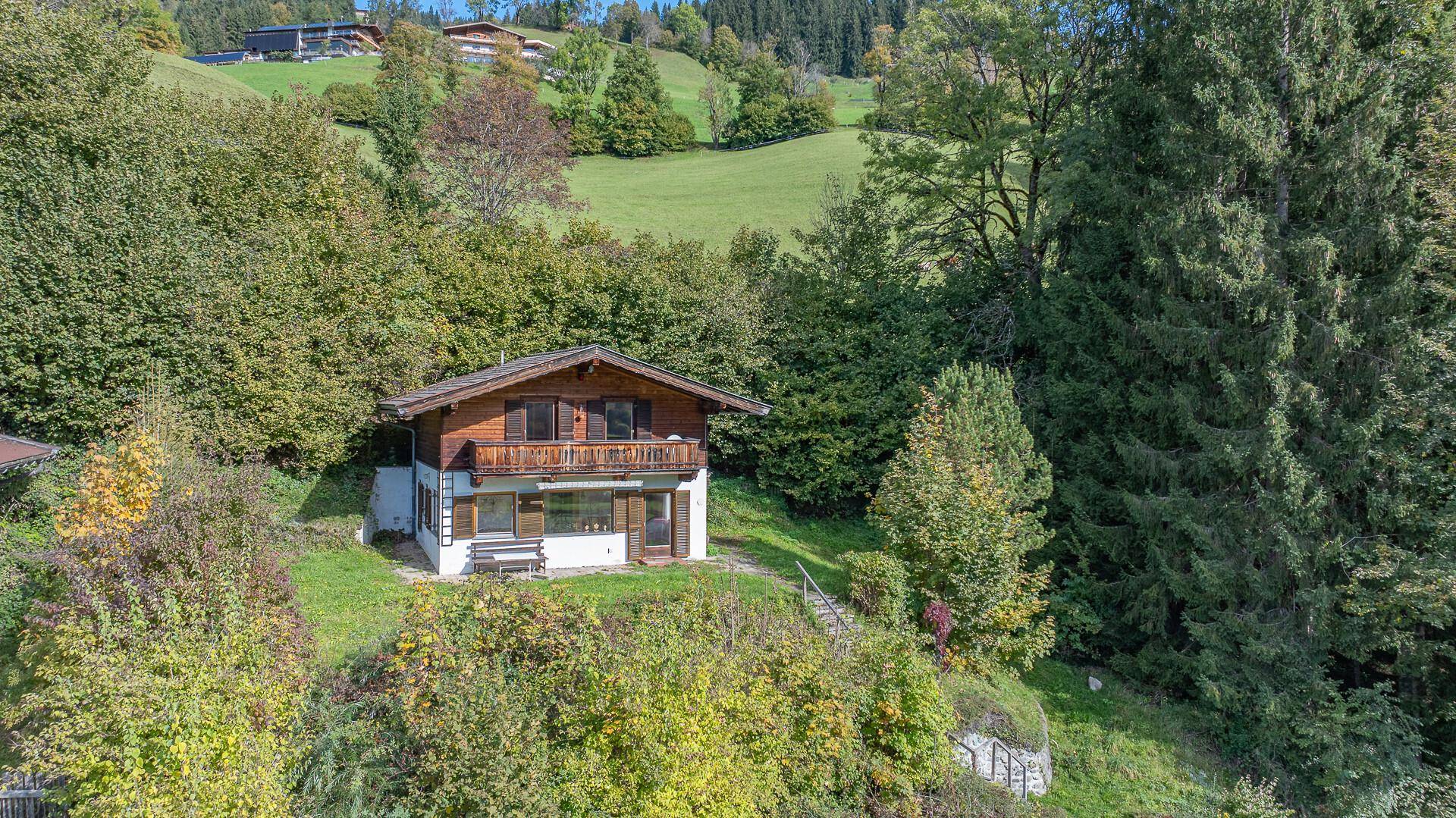 Charmantes Holzhaus mit Balkon, eingebettet in eine grüne Hügellandschaft mit vielen Bäumen.
