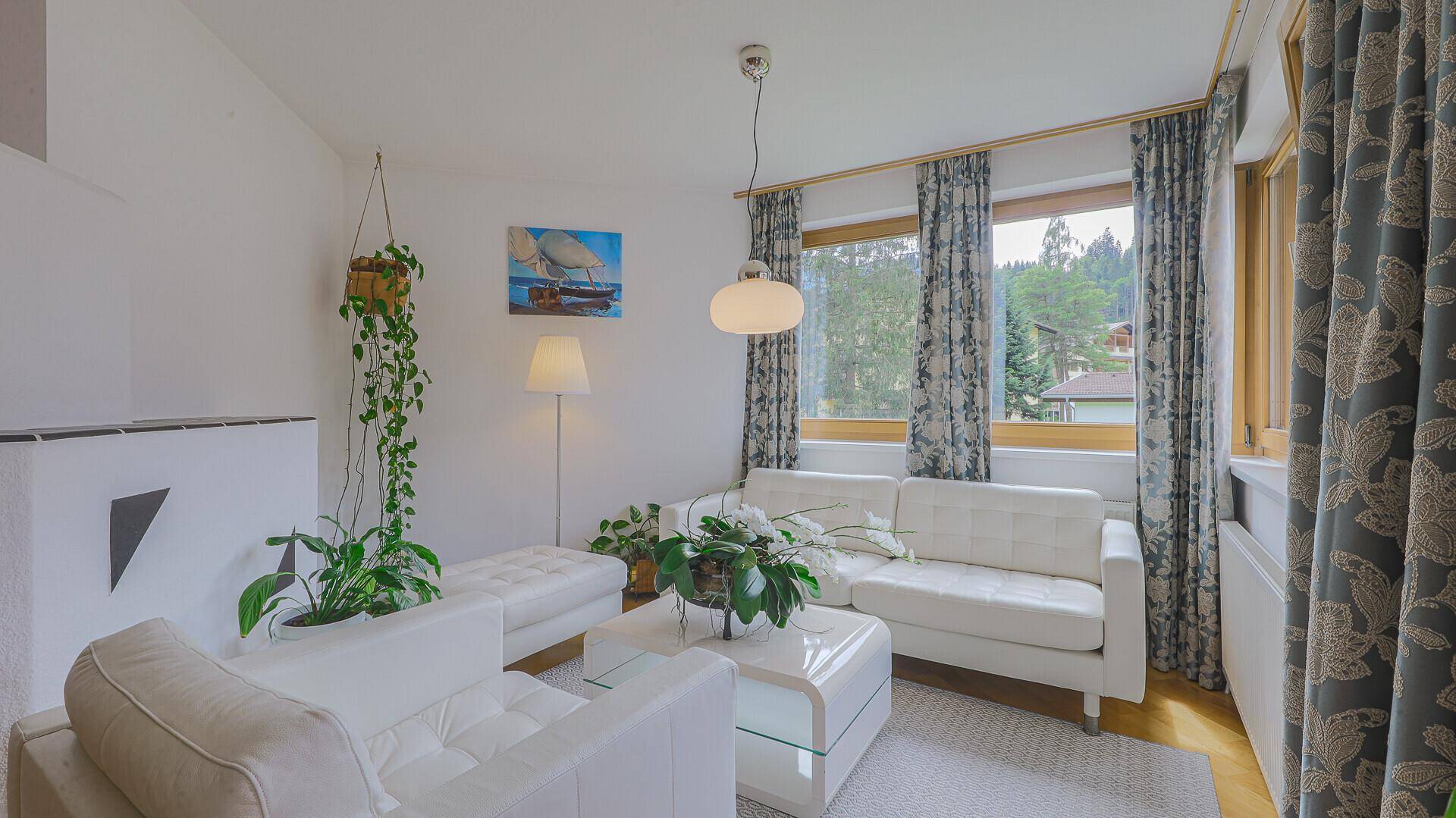Gemütliches Wohnzimmer mit weißem Sofa, großen Fenstern und Blick ins Grüne.