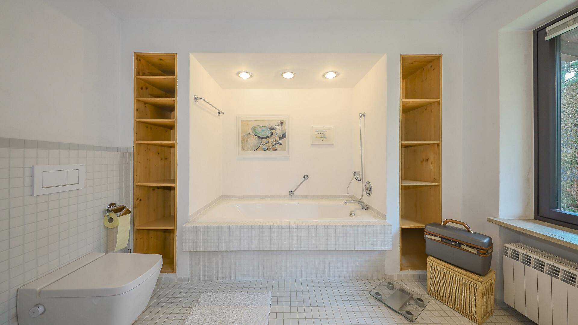 Helles Badezimmer mit eingebauter Badewanne, Holzregalen und Fenster.