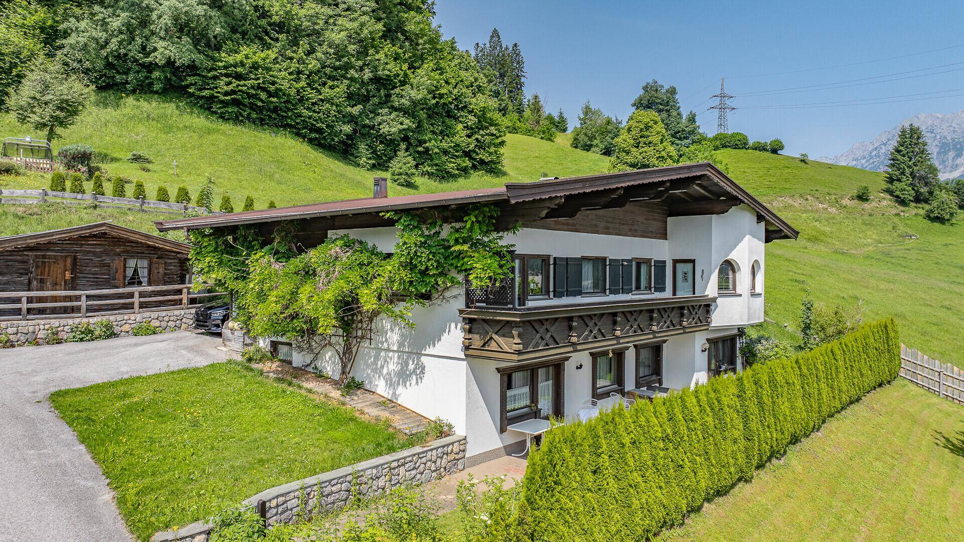 Weißes Haus mit traditionellem Holzbalkon und üppiger Kletterpflanze vor grüner Kulisse.