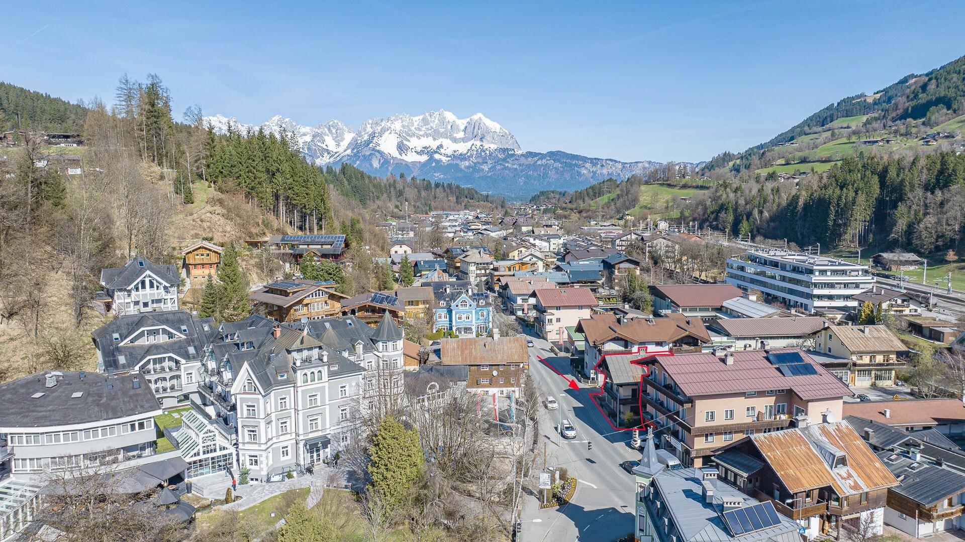 Panoramablick auf die Stadt und die umliegende Bergwelt mit schneebedeckten Gipfeln unter blauem Himmel.