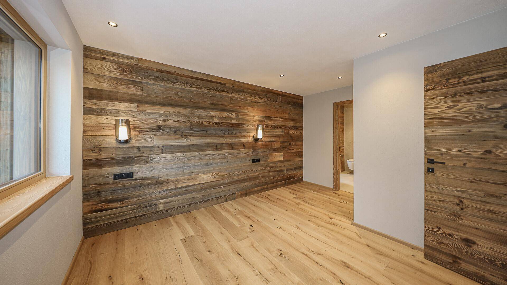 Helles Schlafzimmer mit Holzwand, Holzboden und indirekter Beleuchtung, mit Zugang zum Bad.