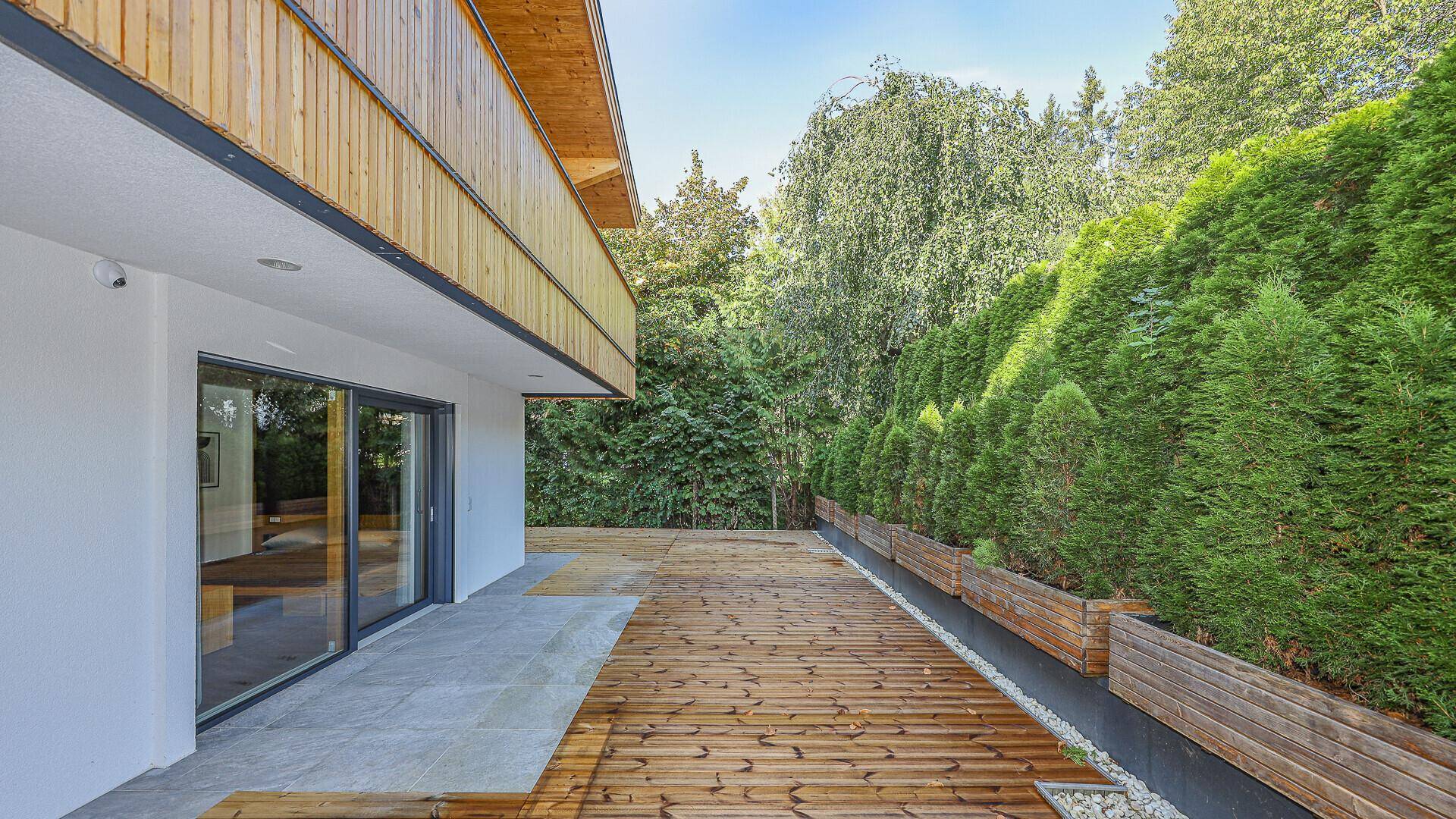 Lange Holzterrasse entlang des Gebäudes mit hoher Hecke für Privatsphäre und Grünblick.