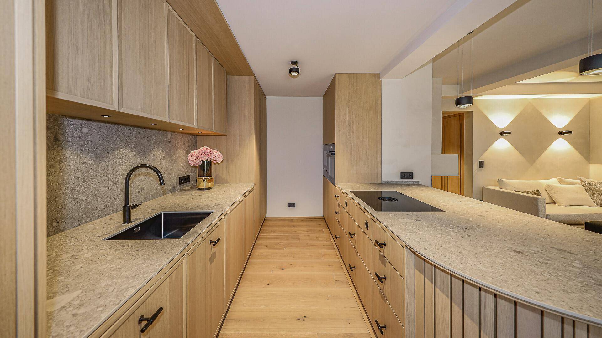 Elegante Holzküche mit Stein-Arbeitsplatten, integrierten Geräten und Blick in den Wohnbereich.