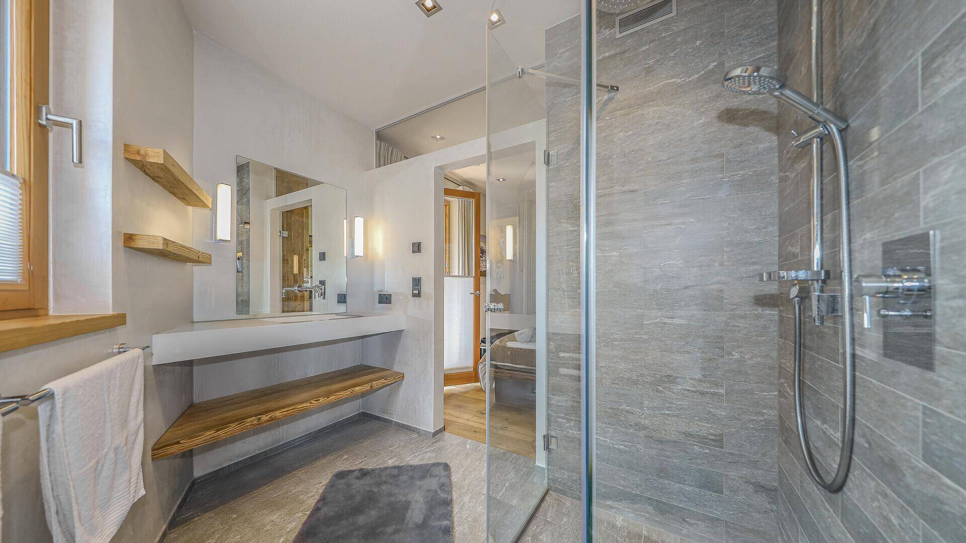 Modernes Badezimmer mit begehbarer Dusche, grauen Fliesen und Holzablage.