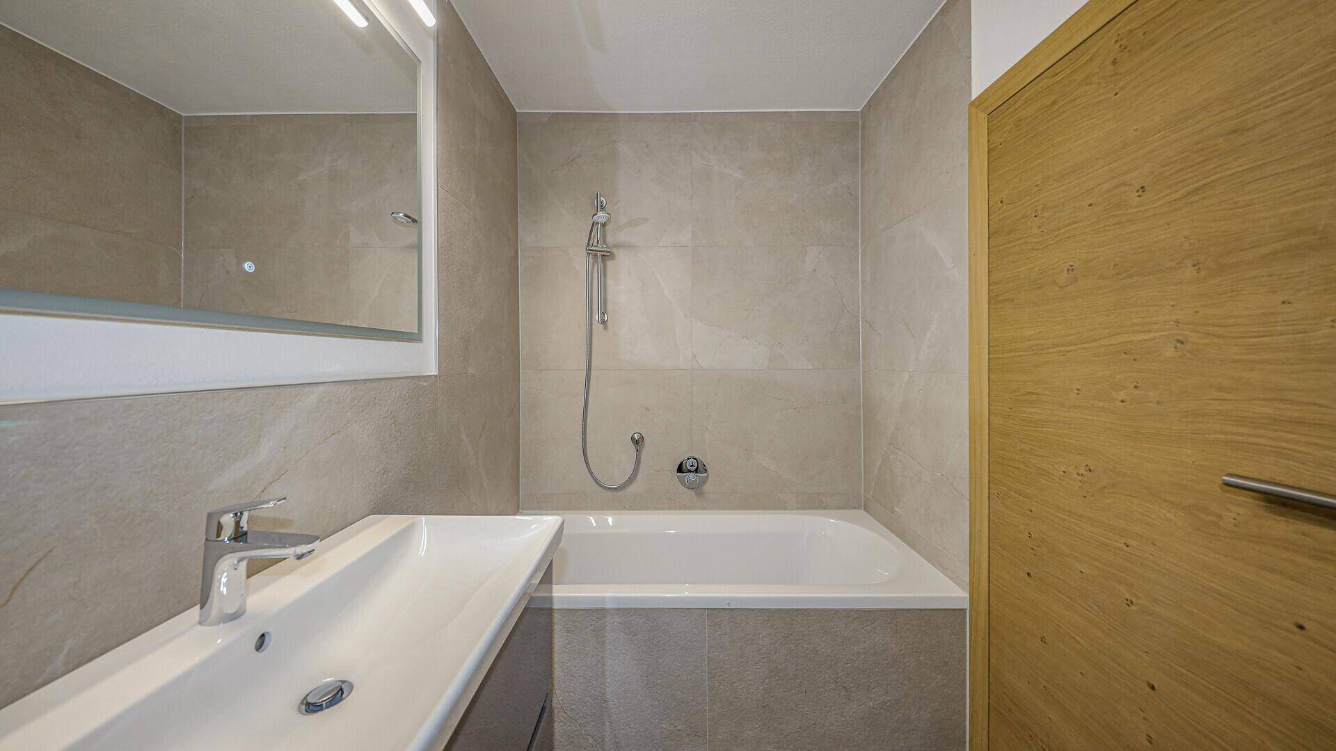 Helles Badezimmer mit grauen Fliesen, modernem Waschtisch und Badewanne mit Duschfunktion.