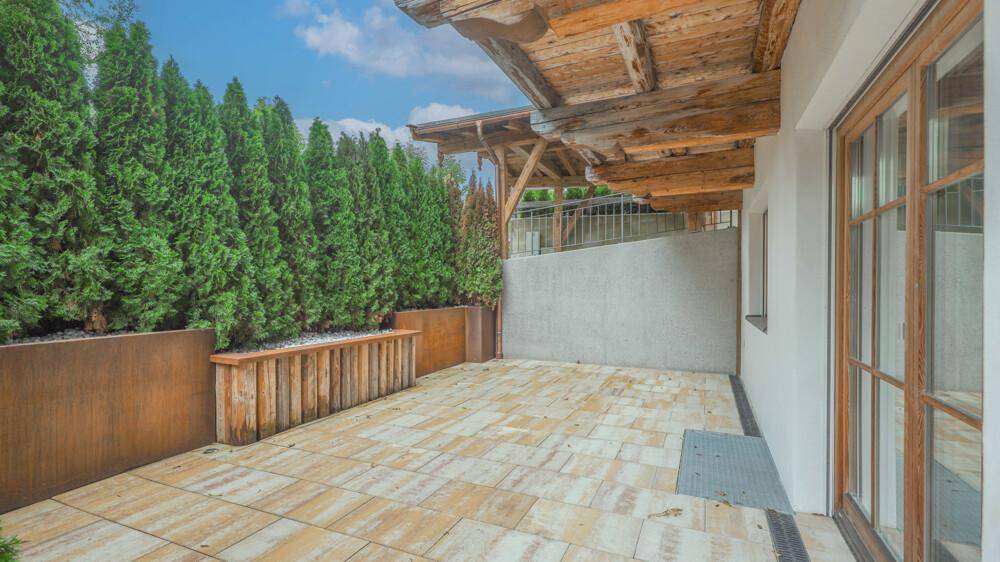 Sonniger Terrassenbereich mit Hecke, Holzpflanzkübeln und gepflastertem Boden.