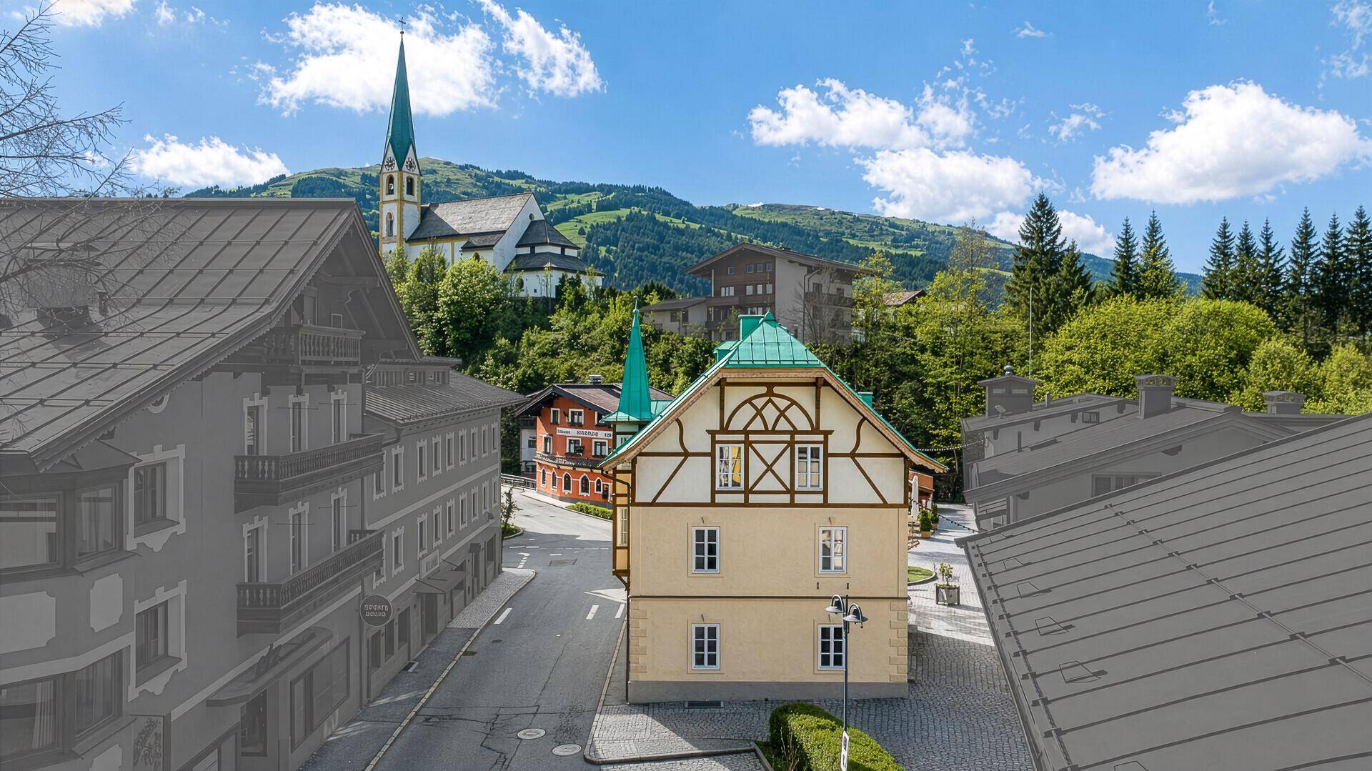 Traditionelles Gebäude im alpinen Stil mit grünem Dach und Fachwerkelementen in einer Berglandschaft mit Kirche.