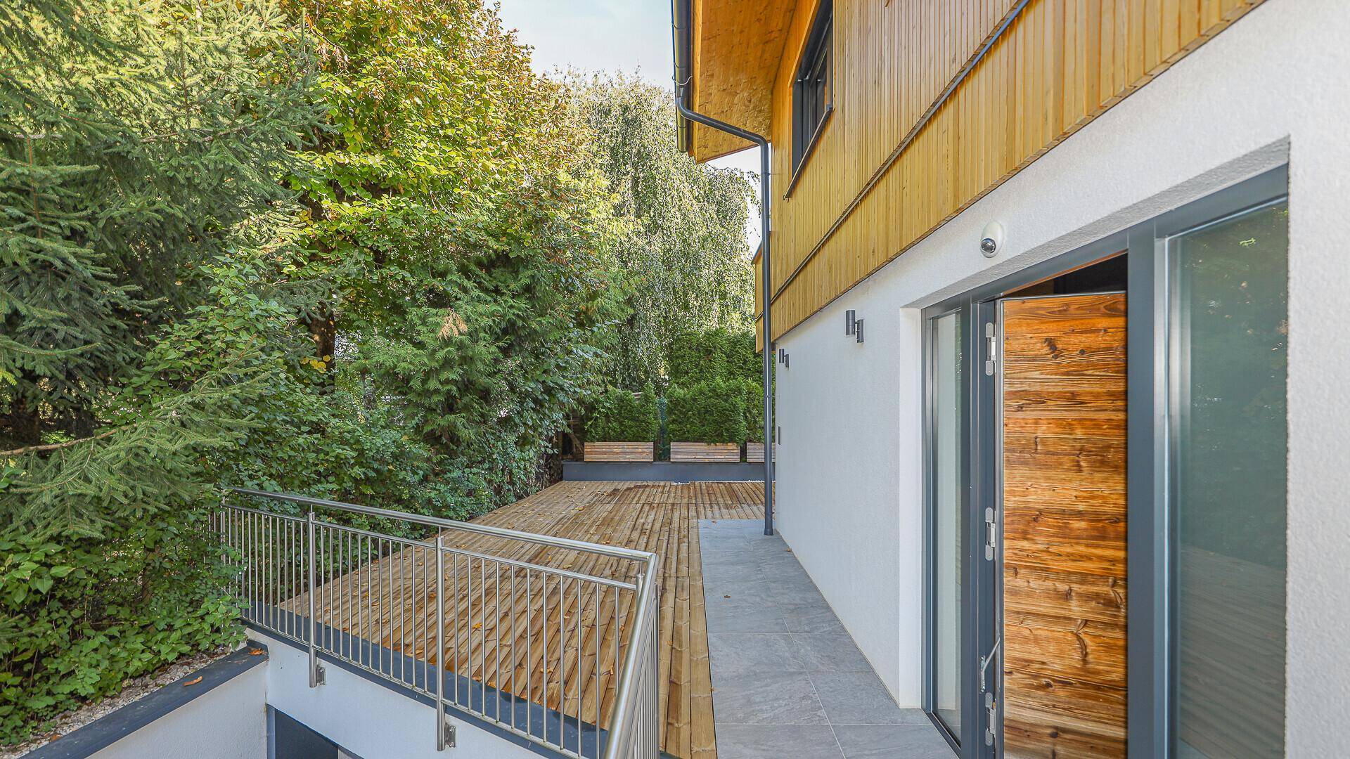 Seitlicher Zugang zur Holzterrasse mit Metallgeländer und umgebender Bepflanzung.
