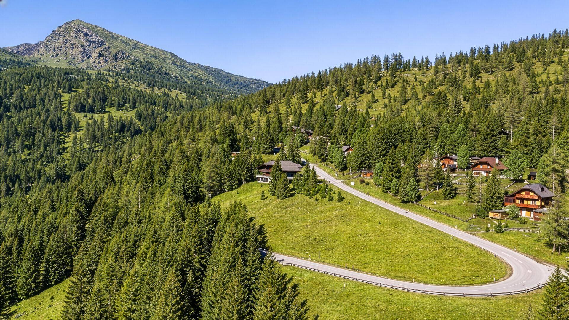 Weitläufige Luftaufnahme einer kurvigen Straße durch eine grüne, bewaldete Berglandschaft mit vereinzelten Häusern.