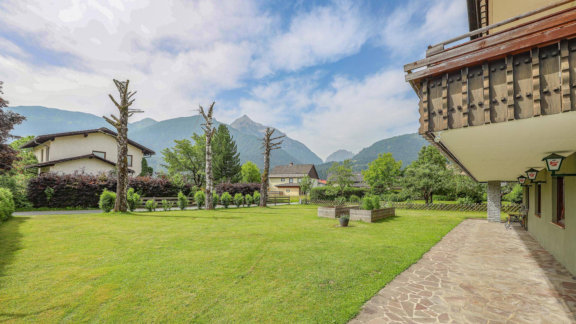 Großer grüner Gartenbereich mit Hochbeeten und einem gepflasterten Weg, umgeben von Natur und Bergen.