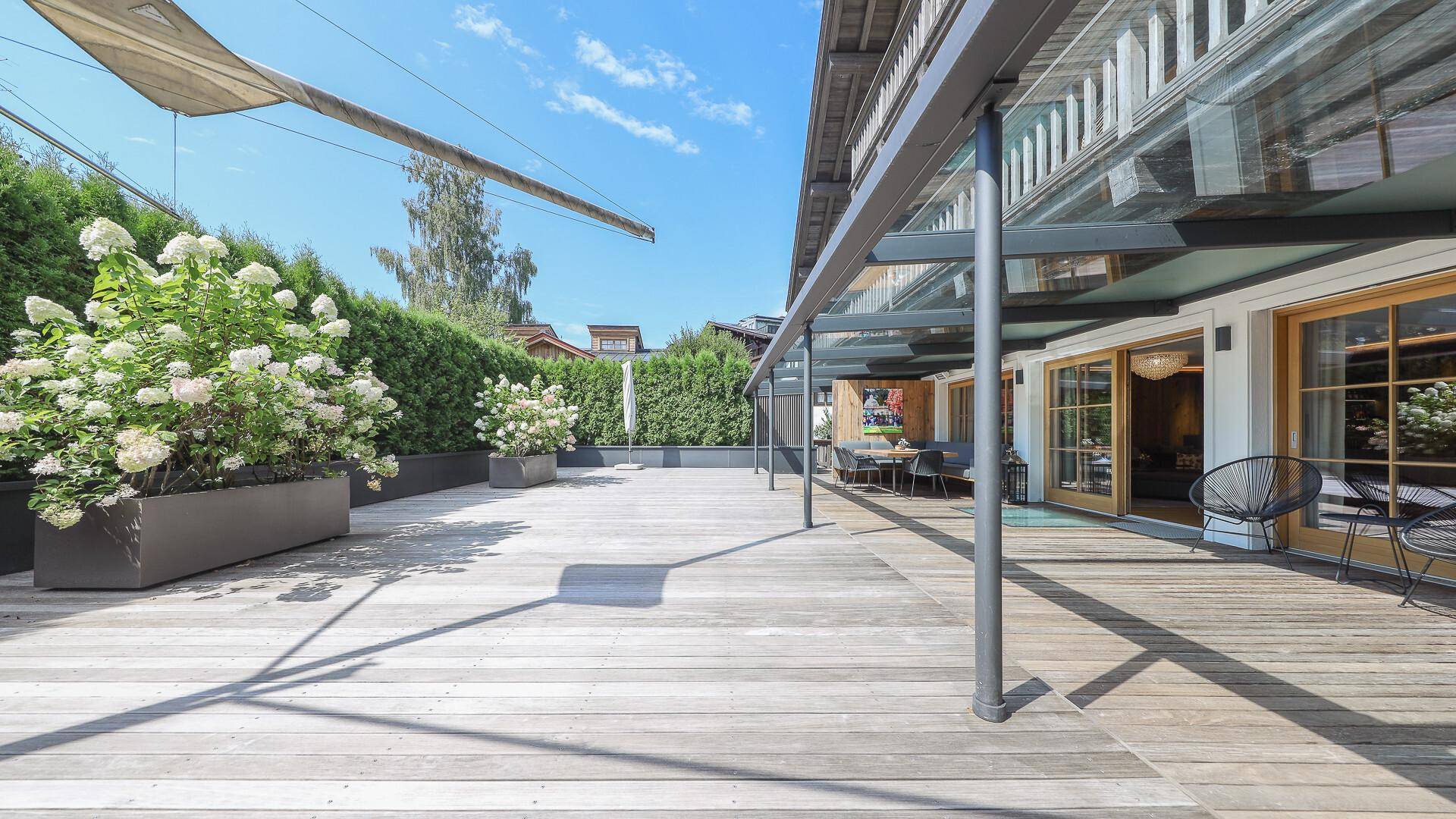 Weitläufige Holzterrasse mit Glasüberdachung, umgeben von grünen Pflanzen und Hecken.