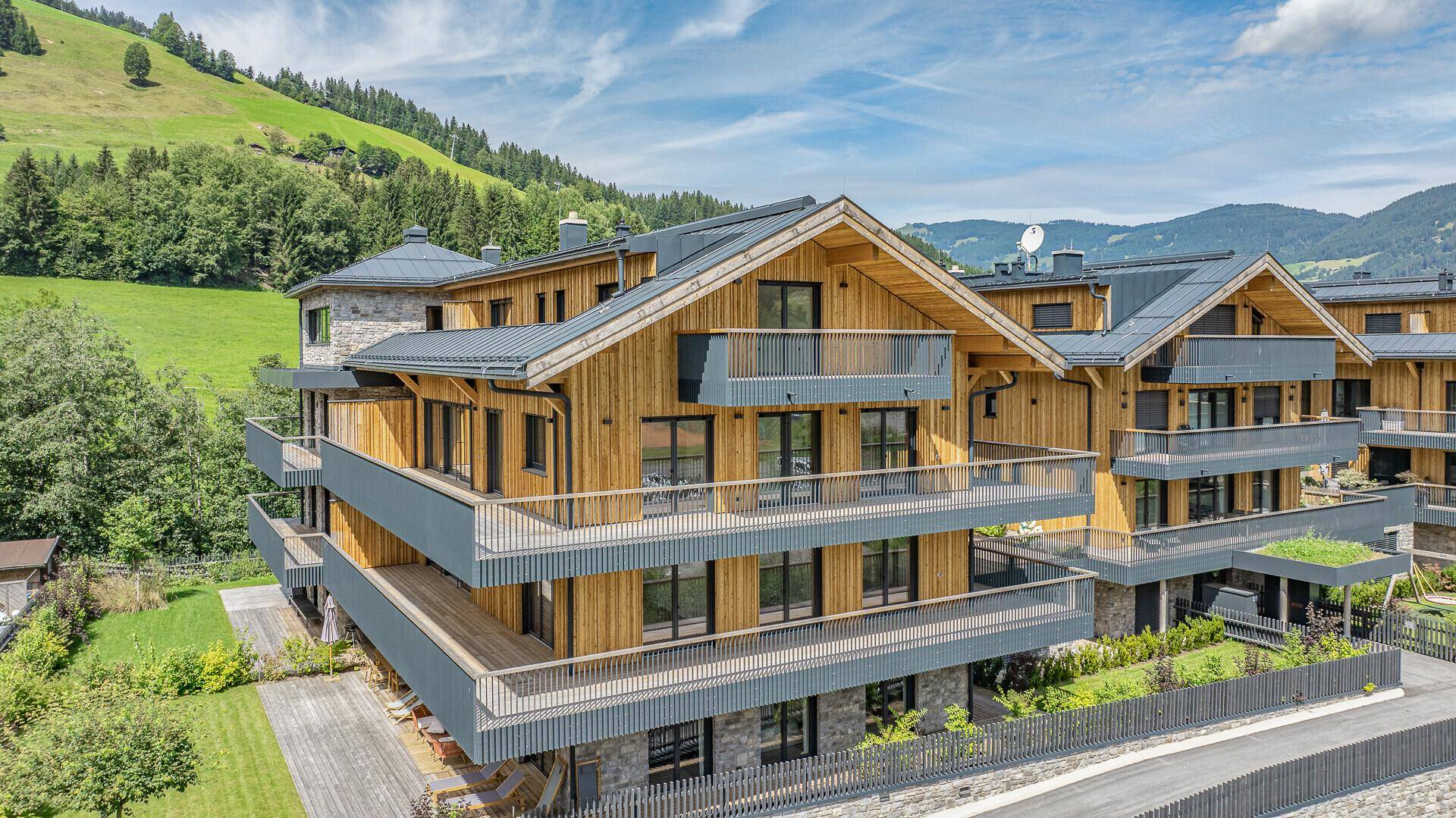 Modernes Apartmentgebäude im alpinen Stil mit Holzfassade und Balkonen vor grüner Bergkulisse.