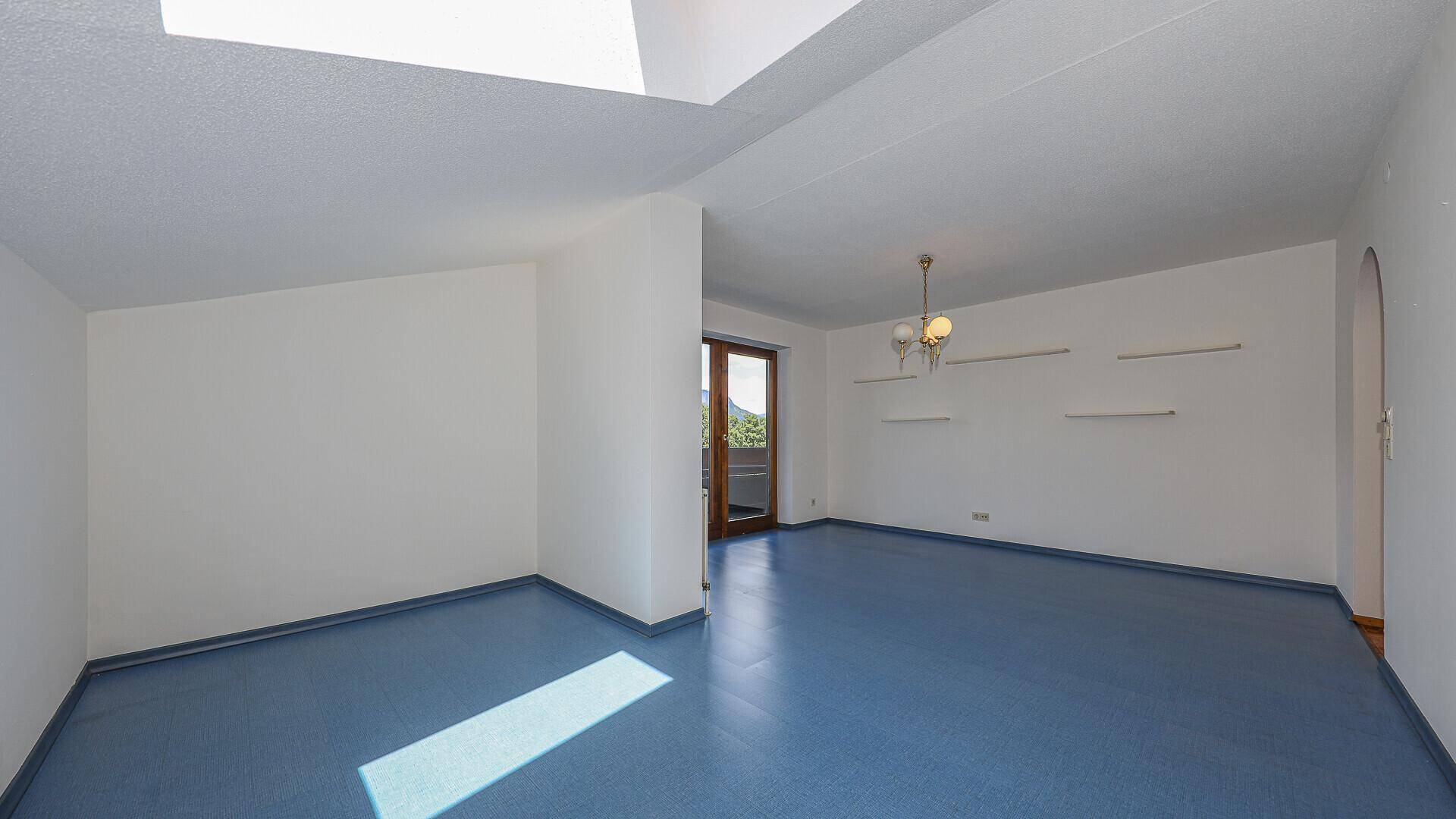 Geräumiger, leerer Wohnbereich mit blauem Boden, Dachfenster und Zugang zu einem weiteren Raum.