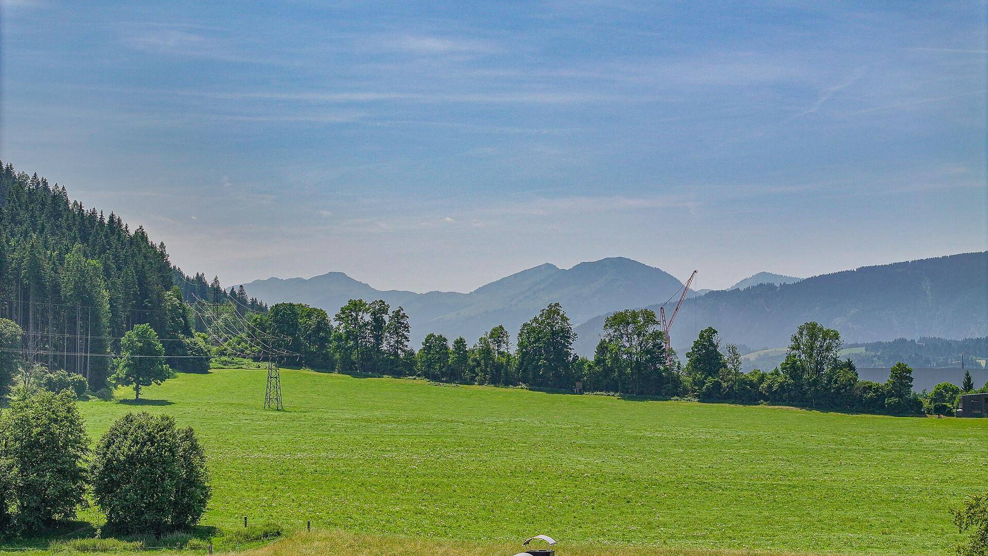 Malerische Landschaft mit grünen Wiesen, Wäldern und fernen Bergen unter blauem Himmel.