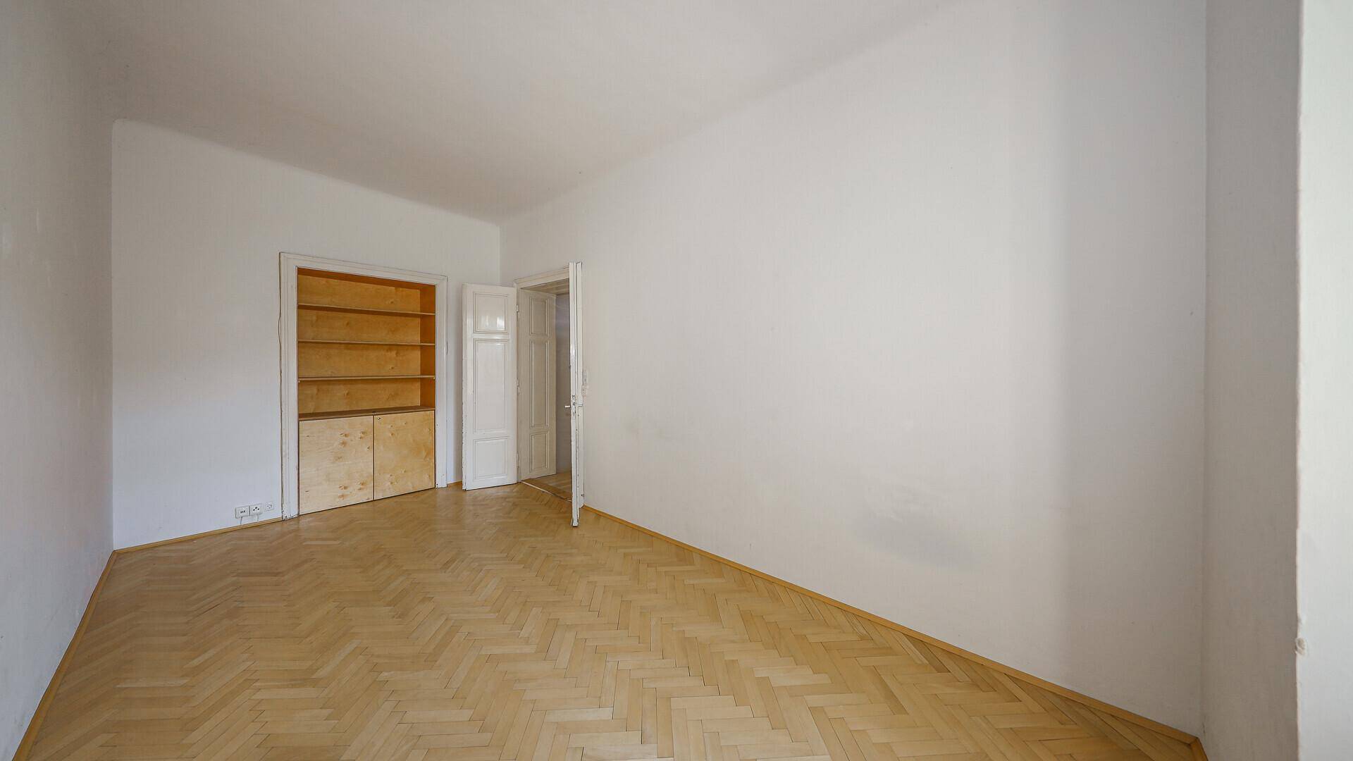 Leeres Zimmer mit Parkettboden, eingebauten Holzregalen und Zugang zu einem weiteren Raum.