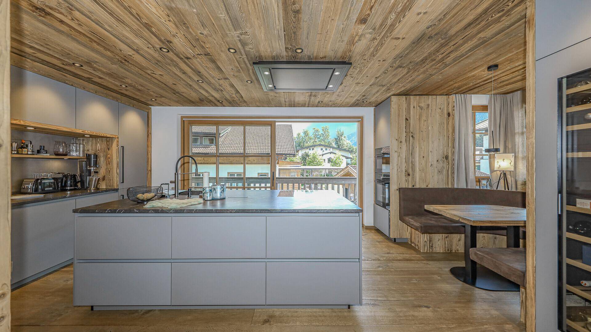 Moderne Küche mit Kücheninsel, grauen Fronten, Holzbalkendecke und Essbereich mit Bergblick.