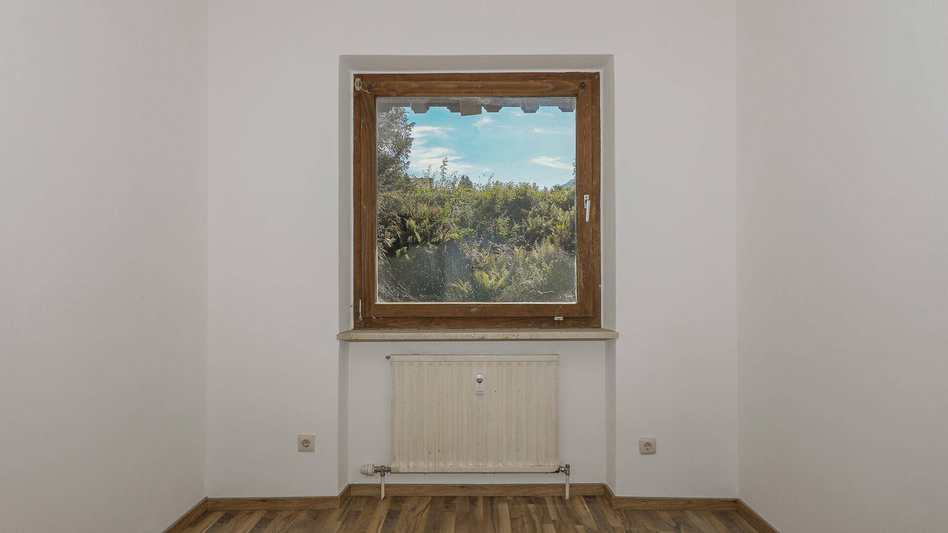 Heller, leerer Raum mit Holzboden, weißen Wänden und einem Fenster mit Blick ins Grüne.