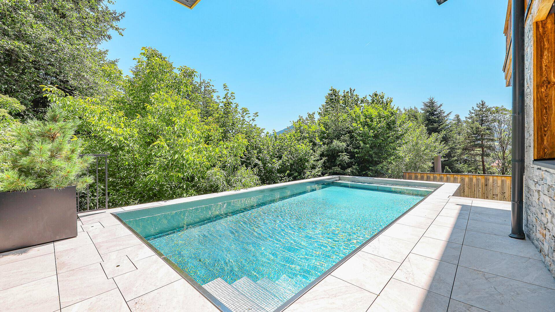 Moderner Außenpool mit gefliester Terrasse, umgeben von grüner Natur unter blauem Himmel.