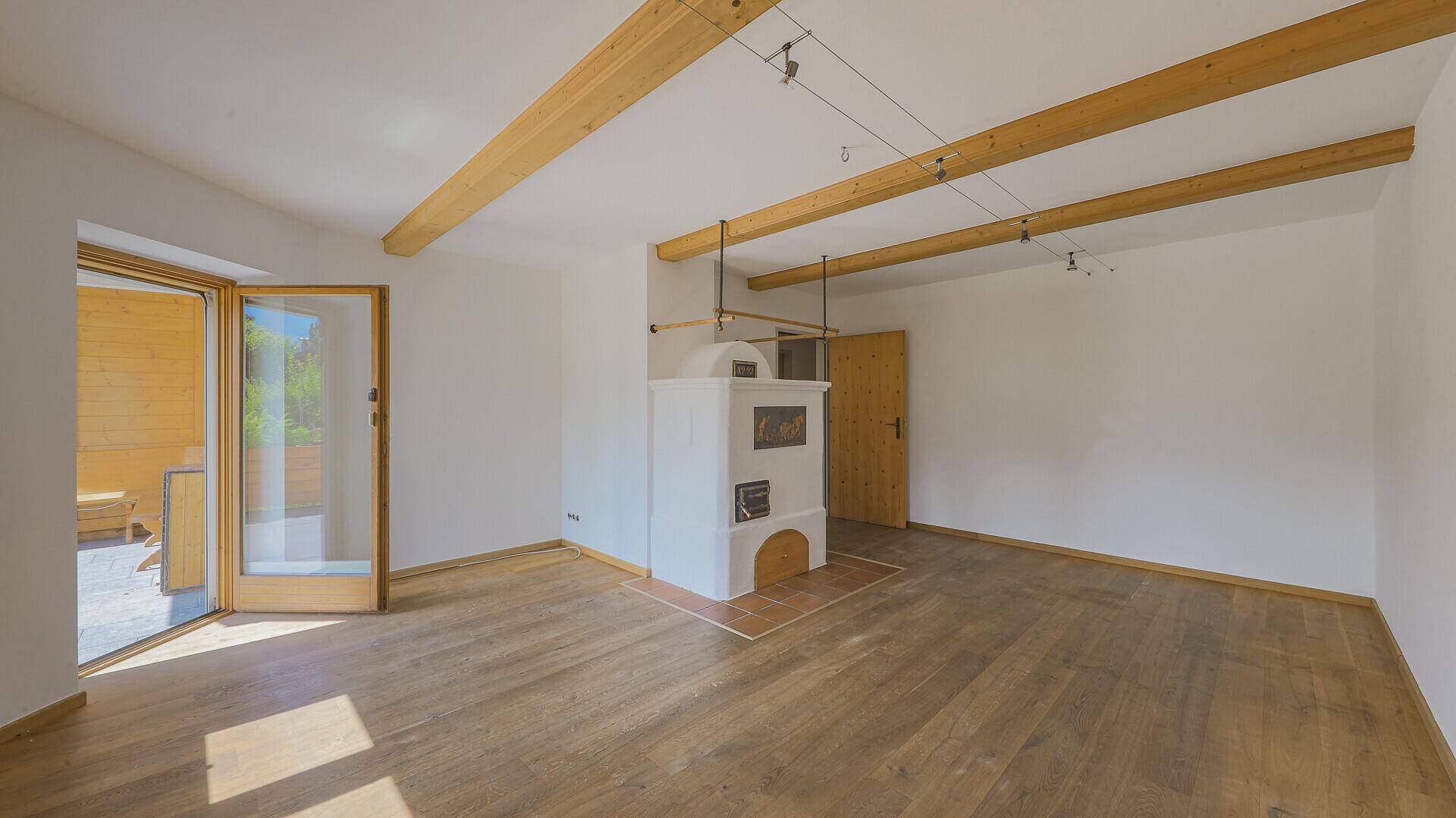 Leeres Wohnzimmer mit traditionellem Kachelofen, sichtbaren Holzbalken und Zugang zur Terrasse.