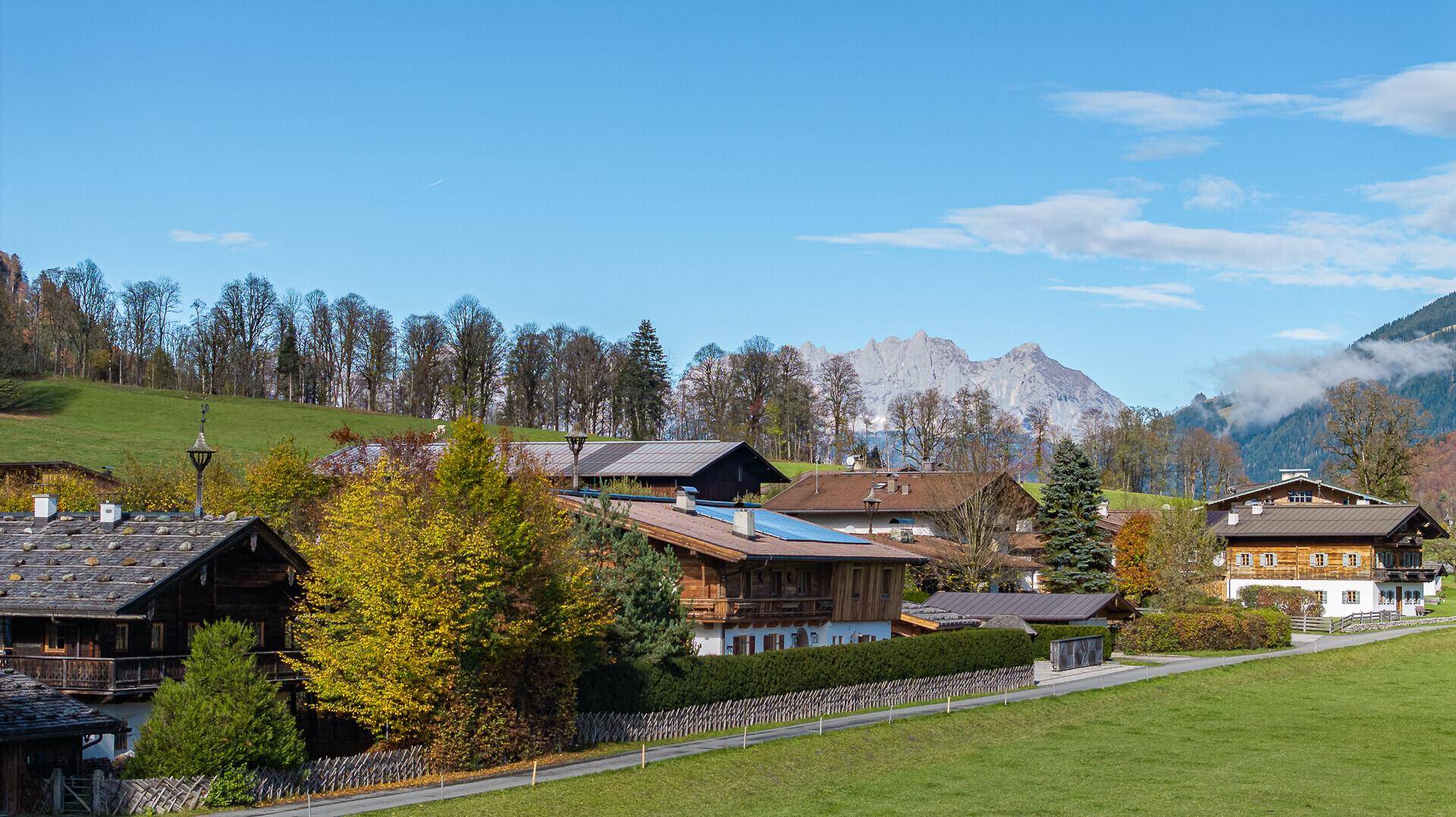 Blick auf traditionelle Häuser in einer ländlichen Umgebung mit grünen Wiesen und Bergen im Hintergrund.