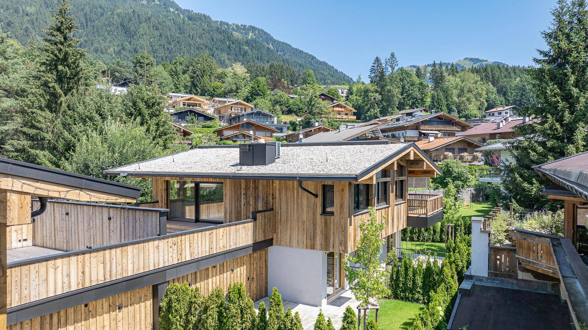 Luftaufnahme eines modernen Holzhauses in alpiner Umgebung mit grünen Gärten und Bergblick.