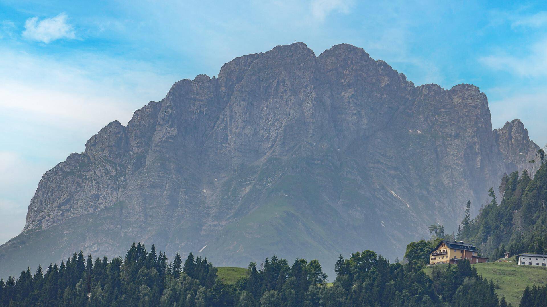 Beeindruckende Bergformation mit einigen Häusern im Vordergrund, umgeben von grünen Wäldern.