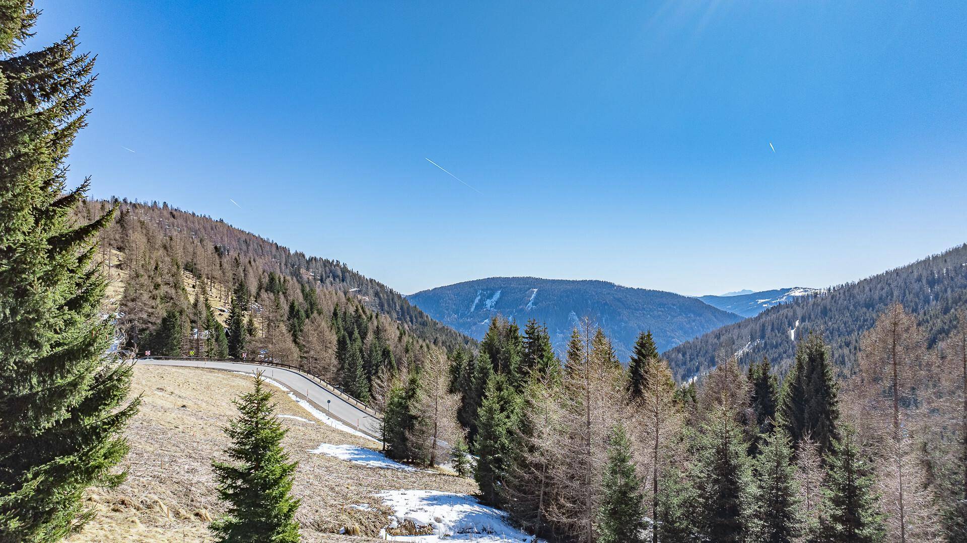Weitläufiger Blick über ein bewaldetes Tal mit einer Straße und klarem blauem Himmel.