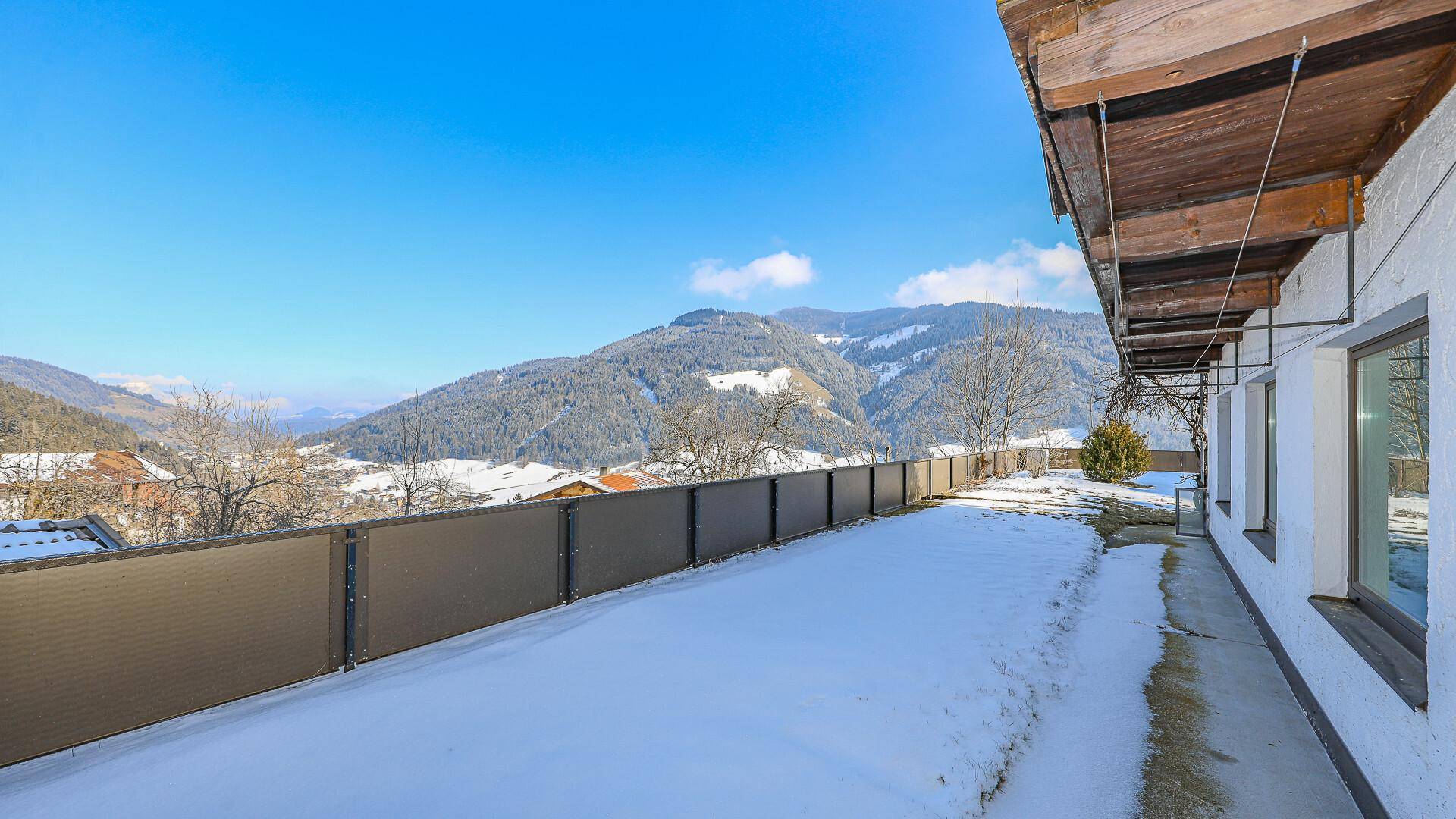 Verschneite Terrasse entlang des Gebäudes mit Blick auf die umliegende Berglandschaft.